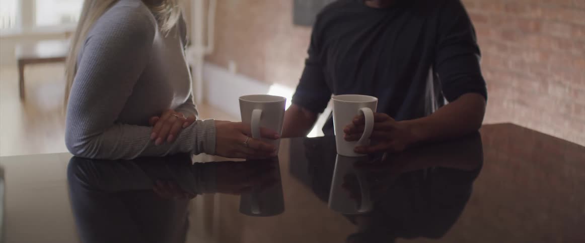 Male and female couple face each other while sitting close together at the kitchen counter while each is holding a coffee mug