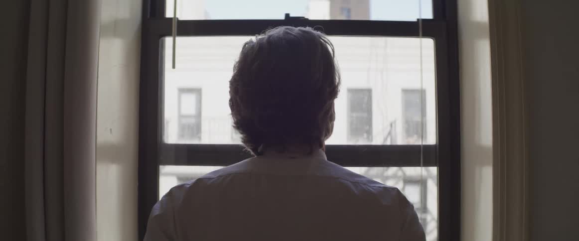 A man stands at a small window looking out at the apartment building across the way