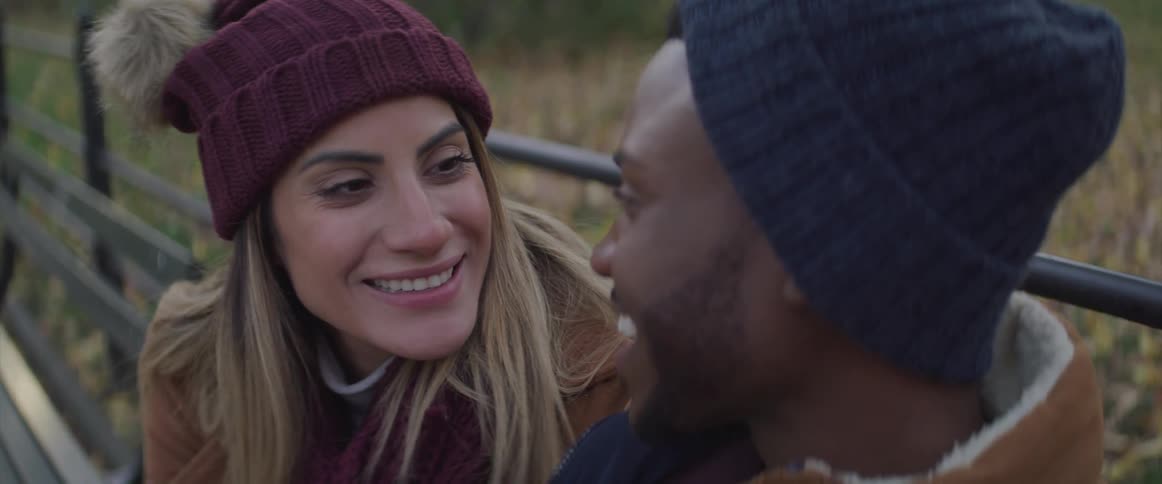 Couple sits closely together on a park bench gazing lovingly into each others eyes while both have a big smile on their face