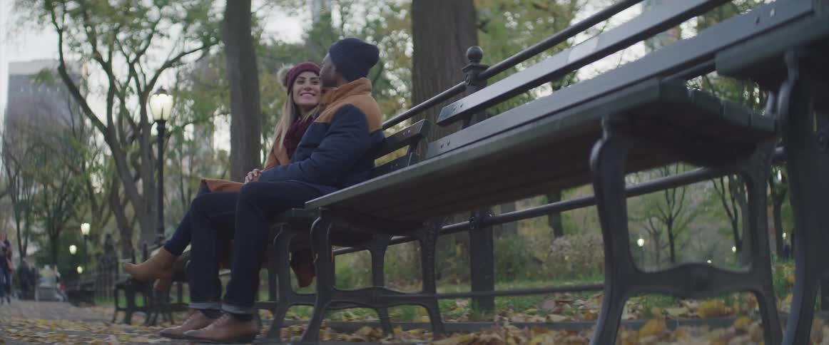 Couple hanging out laughing and talking on park bench at a park in the city