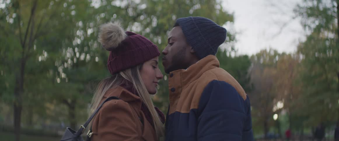 A man kisses his girlfriend on the forehead as they stand in a park
