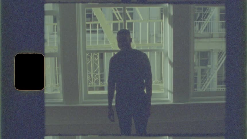 The black man standing in front of the window looks at the camera and laughs
