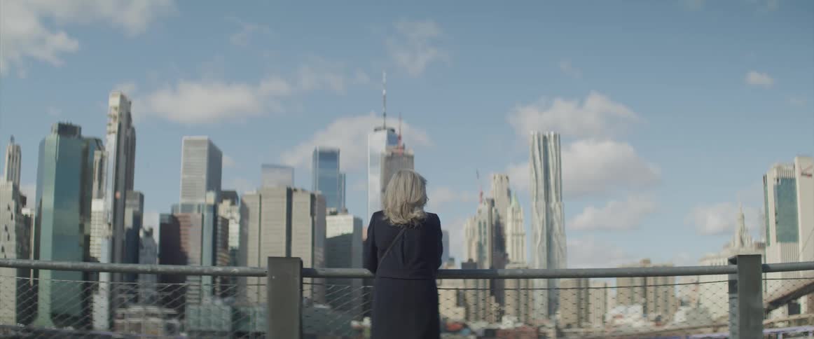 A woman standing outdoors resting her arms on a metal handrail while looking out at the distant skyscrapers of the city