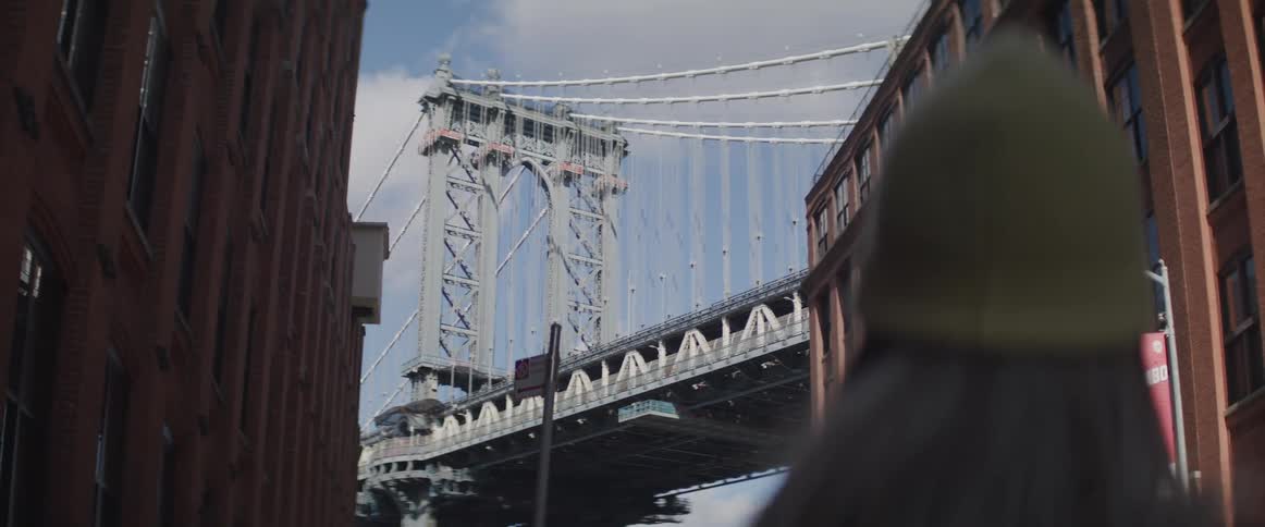A person wearing a beanie hat stands on the street looking up at a bridge on a sunny day