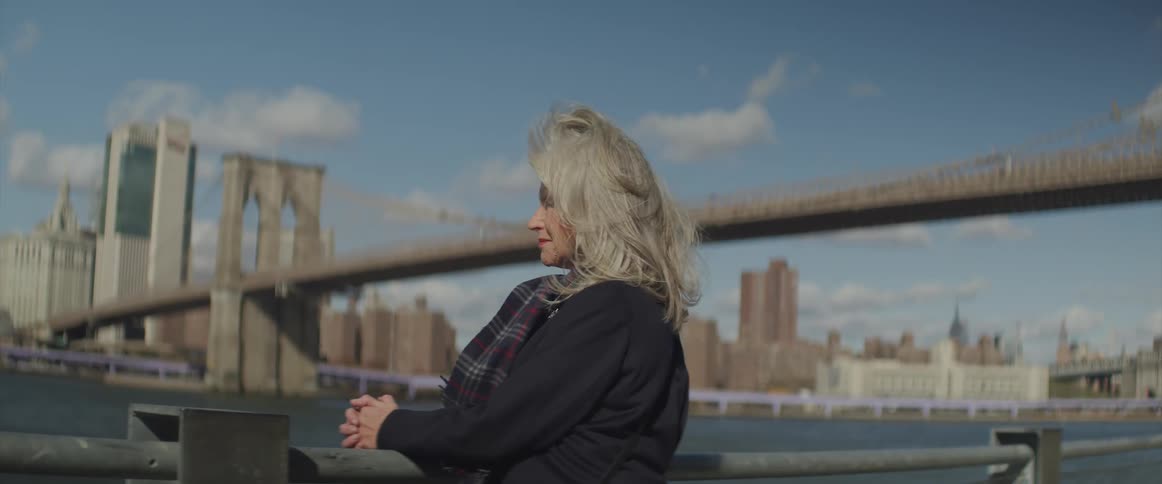A woman stands by a railing next to a river bridge and large city