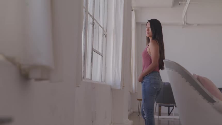 Asian woman stands and looks out the window in the living room at home
