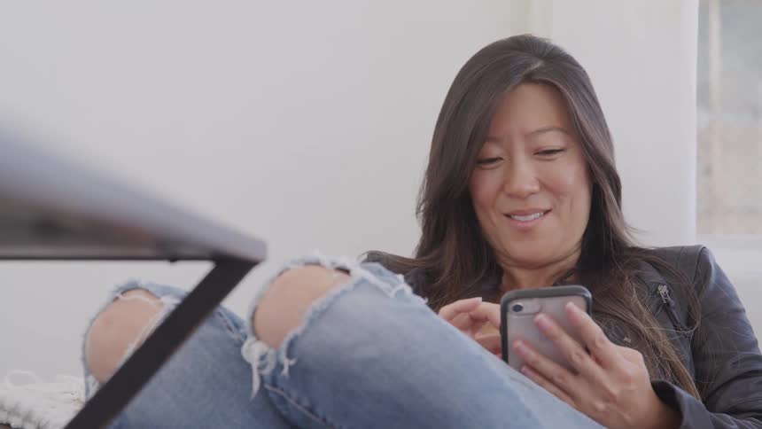 Woman sitting in couch smiles while scrolling on her smart phone