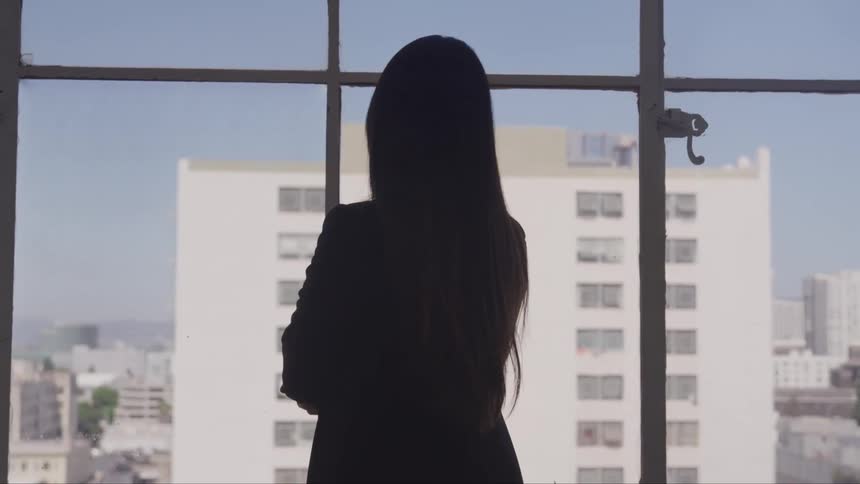 A woman stands in front of floor to ceiling windows looking outside at a tall building