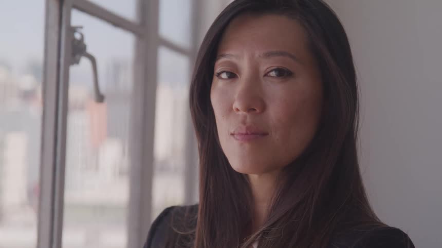 A powerful looking woman stands by a window in a city office and turns her head