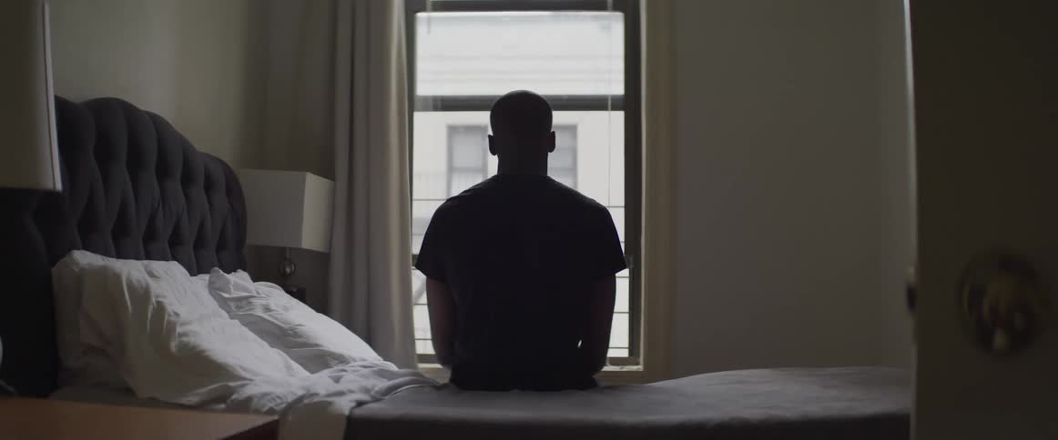 Back shot of a man facing a window while sitting on a bed possible in a hotel room