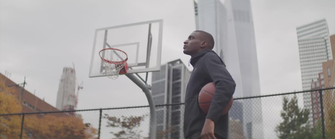 Male holds a basketball under his arm as he stands on a basketball court in the city and looks up in the air with a serious look on his face