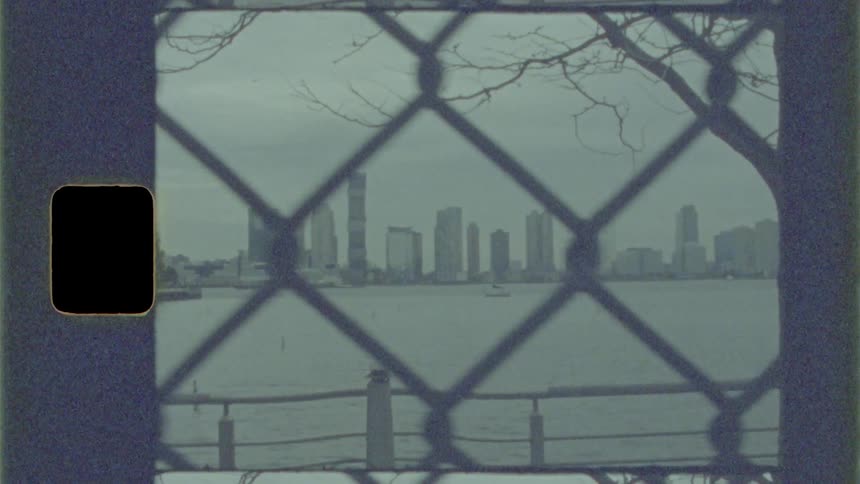 Through a chain link fence a city skyline is seen across the ocean