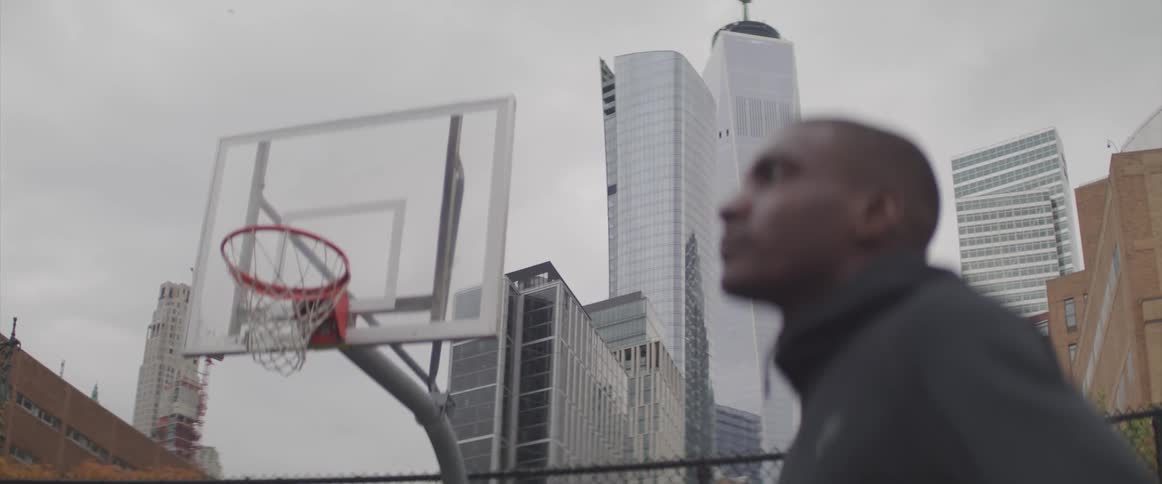 A man stands in profile on an outdoor basketball court with the hoop and city buildings behind him on a cloudy day
