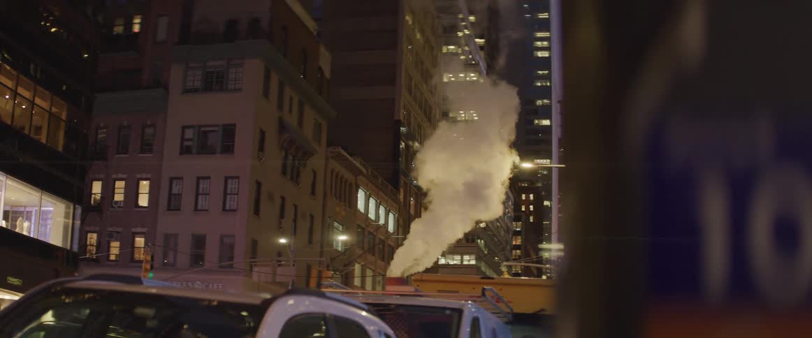 A city street with cars parked and steam rising out of a stack