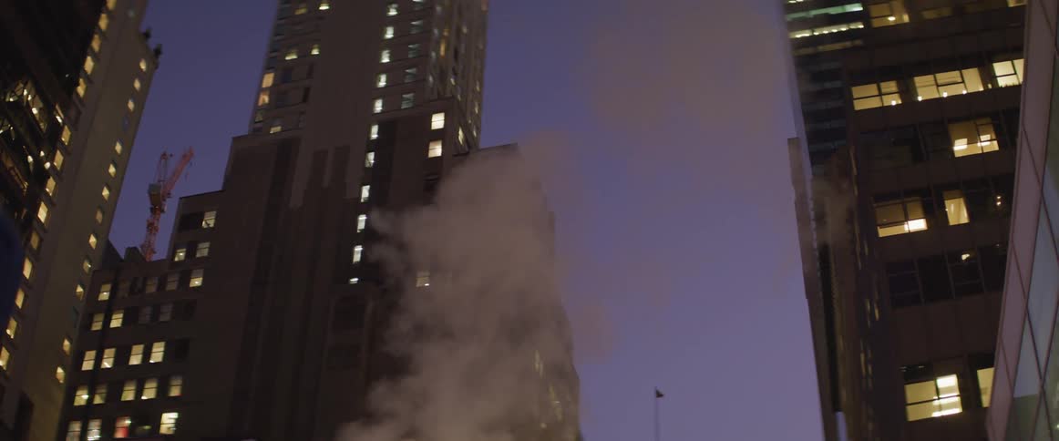 Light smoke rises from the ground in front of tall skyscrapers at night
