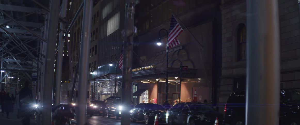 People walk on a covered sidewalk with cars parked and stopped on a city street at night