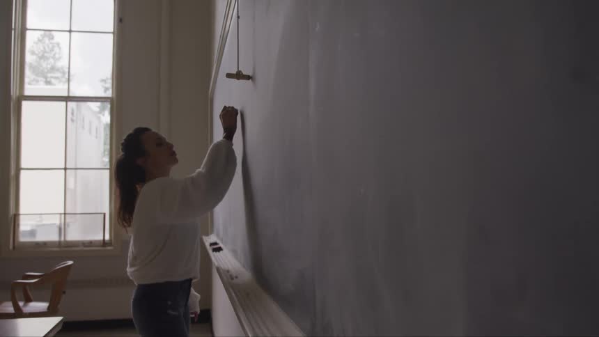 Female teacher carefully writes on the chalkboard in a classroom
