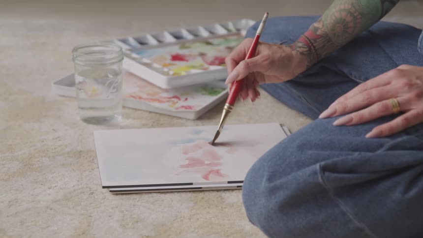 A white woman sits on the floor and paints with watercolors
