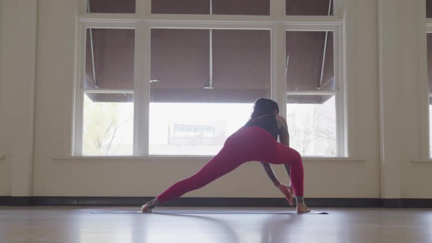 A woman stands in front of a large window stretching during a yoga exercise