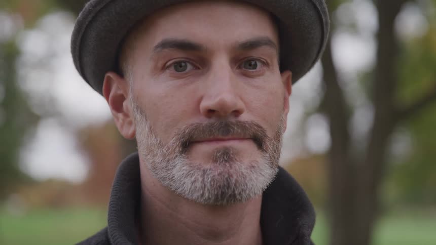 Bearded man wearing a hat stands and looks at the camera in the public park