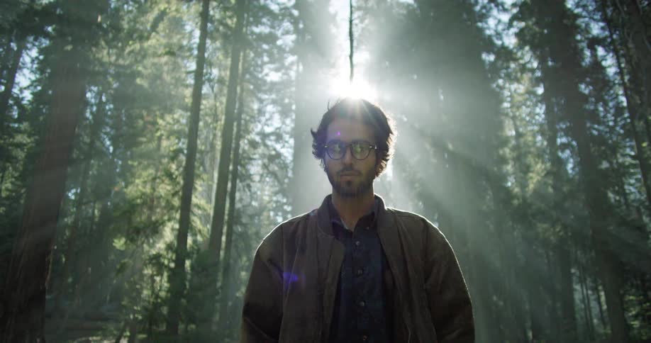 A white man stands in a forest as the sun shines down
