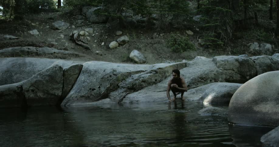 A man crouching down in front of a pond and splashing water on his hands