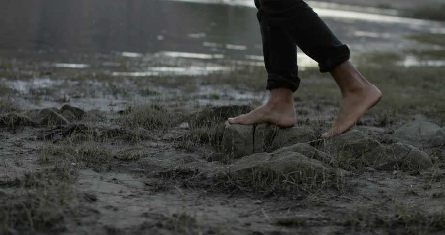 A man with pants rolled up and bare feet walks on rocks and sand heading to the lake