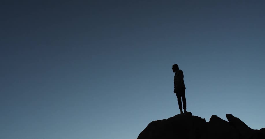 Silhouette of a person standing on peak of large rock against blue skies