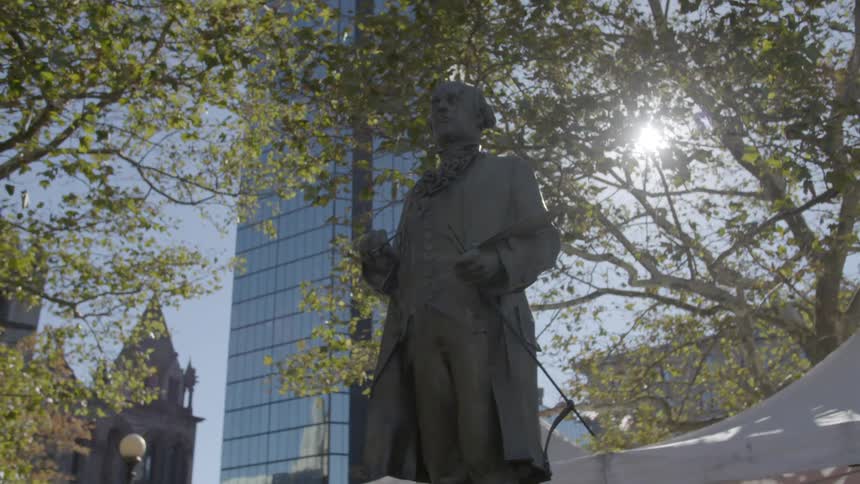 A statue of a man in historical clothing sits in a public park near tall buildings