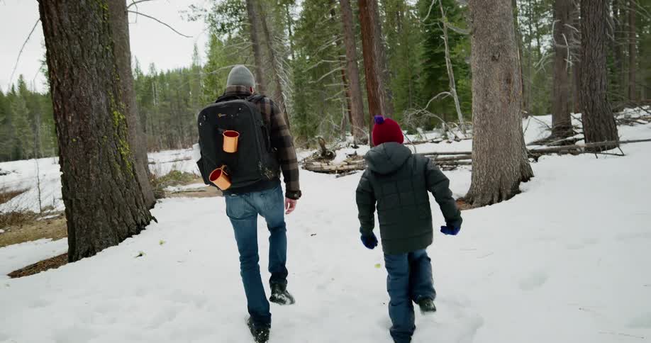 A child and adult walk through a snow covered woods area with a backpack for camping