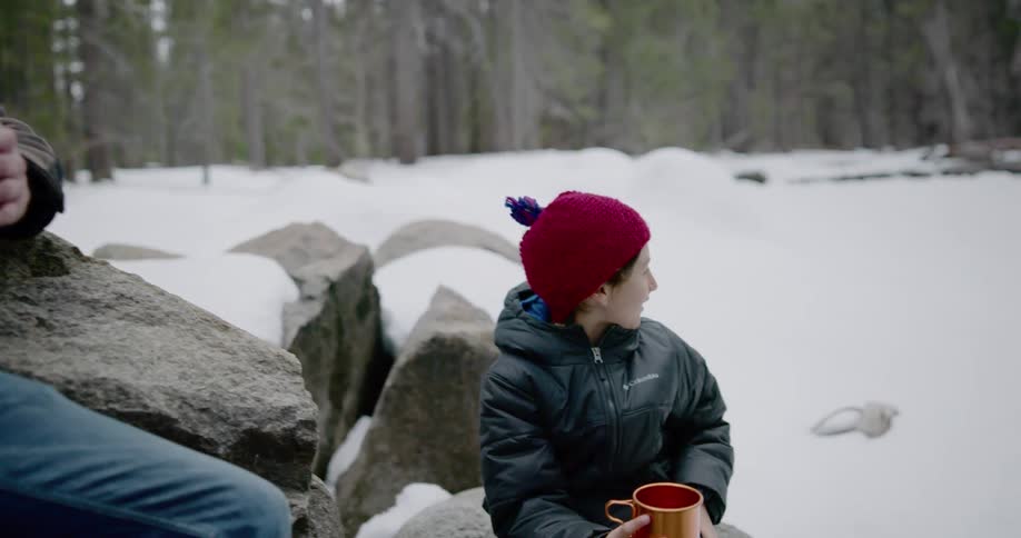 A dad and his child sit on rocks drinking from a thermos in the snow woods talking and pointing