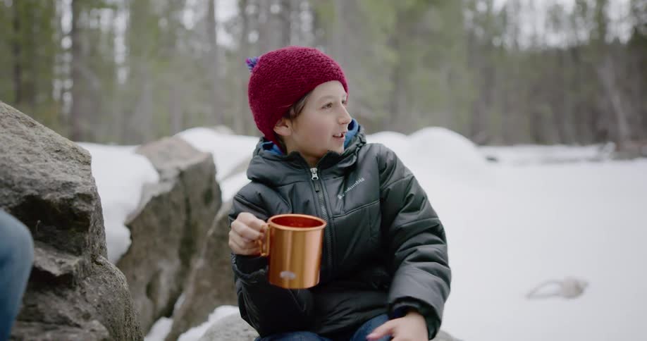 A young boy sits and drinks from a mug outside in the snowy woods and talks to another person