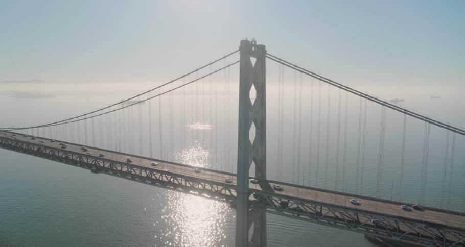 Traffic crosses suspension bridge over water as mist obscures boats on the horizon