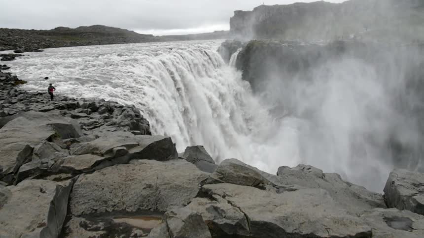 A large waterfall has one person standing on the rocks above