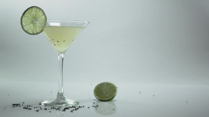 A cocktail in a martini glass garnished with a slice of lime has seeds added to it