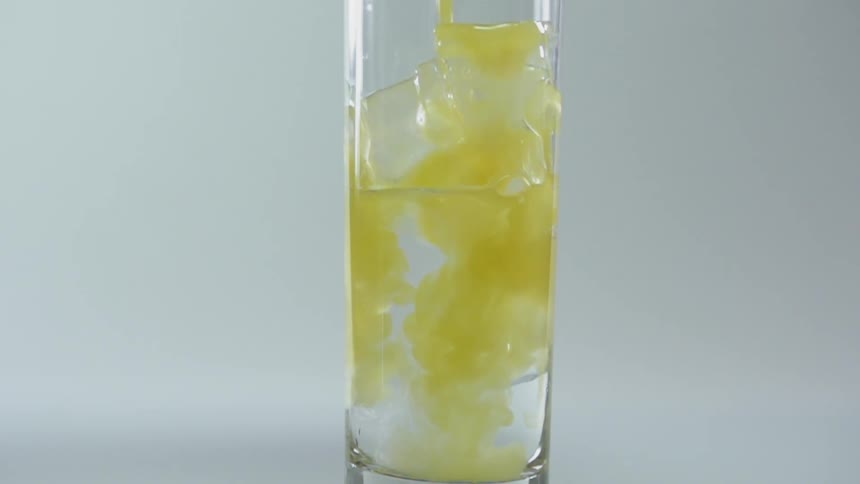 A yellow liquid being poured into a long narrow drinking glass filled with ice