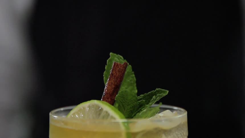 A mint leaf garnish drops onto a cocktail already garnished with a slice of lime and a cinnamon stick