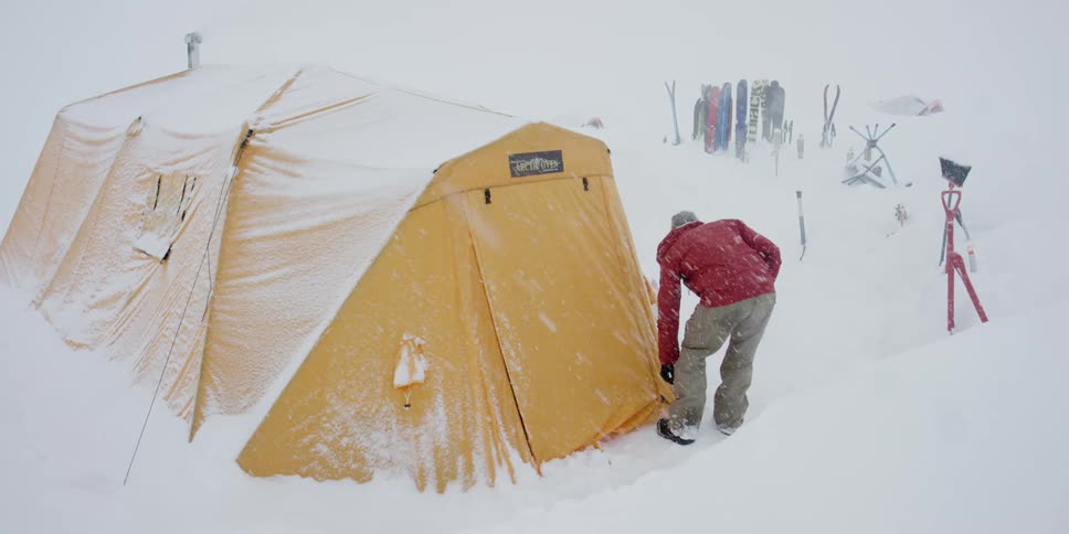 A person with a red jacket closing a yellow tent then walking in the snow