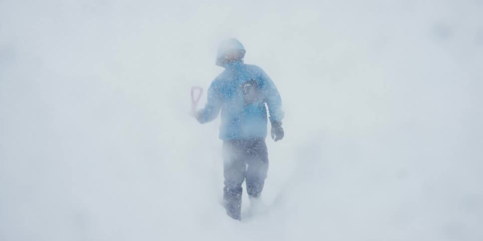A person walking through knee deep snow carrying a shovel as snow falls heavily from the sky