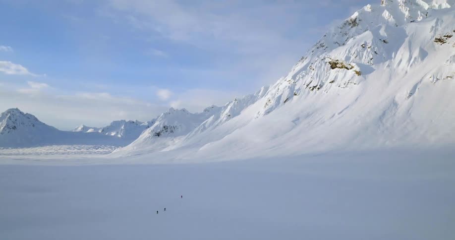 Aerial of people skiing in a snowy valley next to a mountain range covered in snow