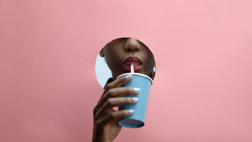 A woman drinks from a cup with a straw in it holding it to her mouth through a round cutout in pink paper