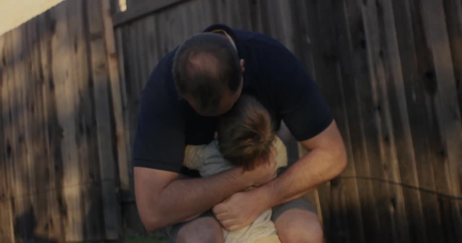 A man in a yard bends down and hugs a child