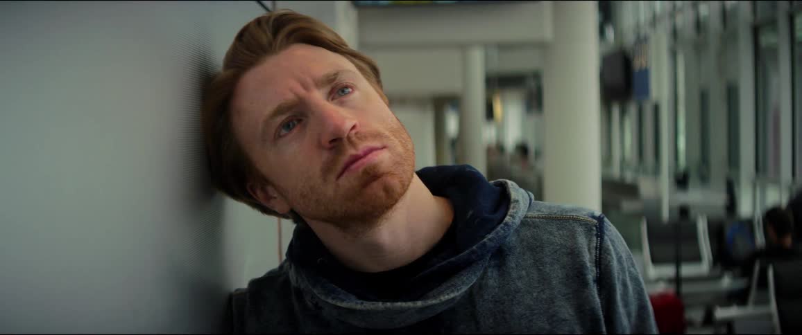A white man leans against a wall inside a building