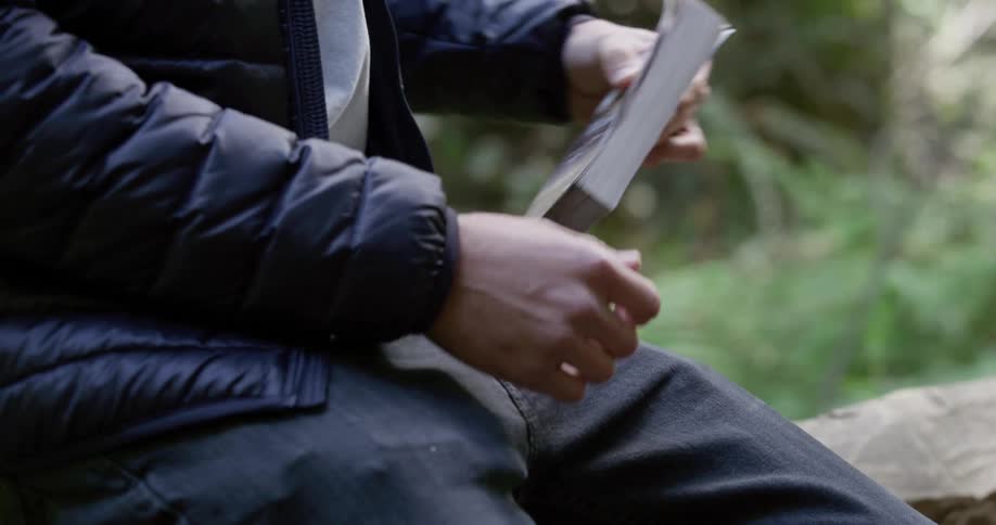 Male sitting on log in the woods removes a book from his back pocket and looks at the front cover while resting his hand on his lap