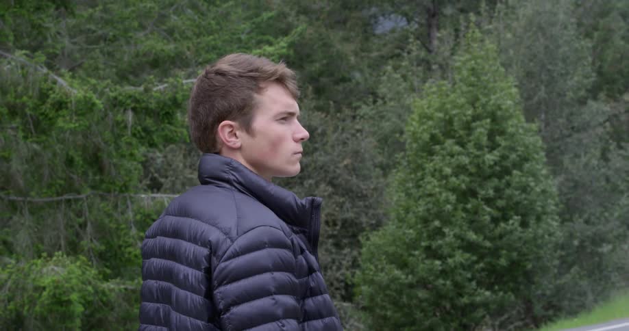 A man stands by a large forest area and road wearing a coat