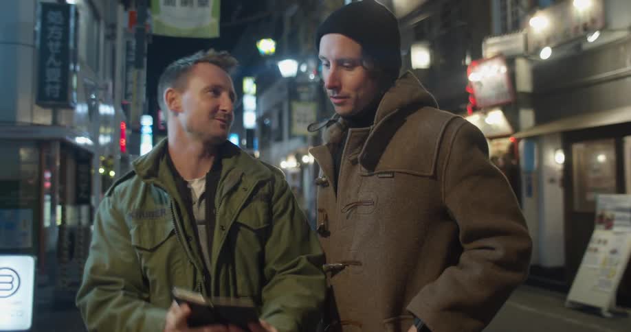Two men stand on the street talking and smiling as they each look at their cell phone