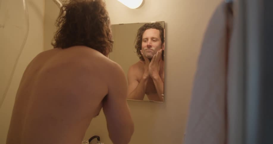 A shirtless man stands at the bathroom sink looking in the mirror and rubbing his face to prepare for shaving