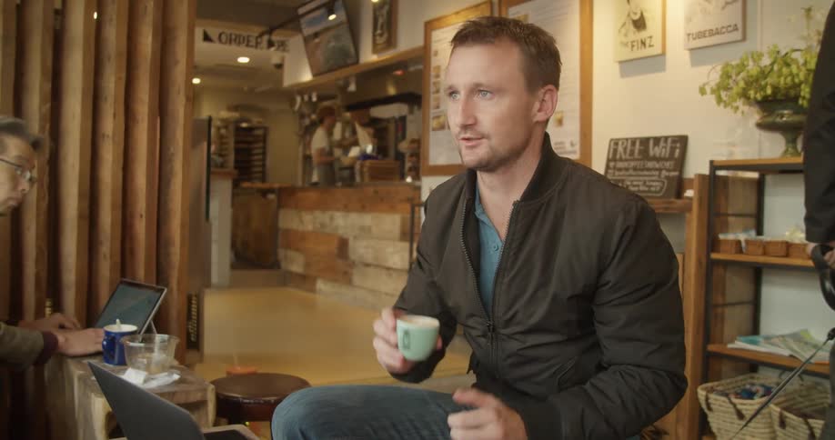 A man sitting in front of a laptop and drinking from a cup in a cafe while talking to another person sitting across from him