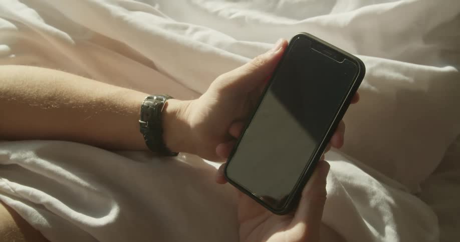 A white male lies in bed while holding a smartphone