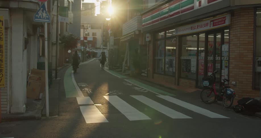 A delivery man pushes a cart towards a town corner store and a woman rides past on a bike